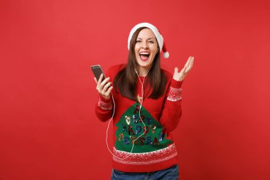 Müzik dinlerken neşeli Santa içindeki genç kız cep telefonu, holding kulaklık ile Noel şapka kırmızı zemin üzerine izole. Mutlu yeni yıl 2019 kutlama tatil parti kavramı. Kopyalama alanı alay