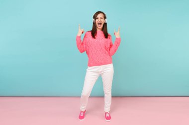 Tam uzunlukta örme gül kazak, beyaz pantolon, parlak pembe mavi pastel duvar arka planda Studio izole kulaklık mutlu genç kadın portresi. Moda yaşam tarzı kavramı. Kopyalama alanı alay