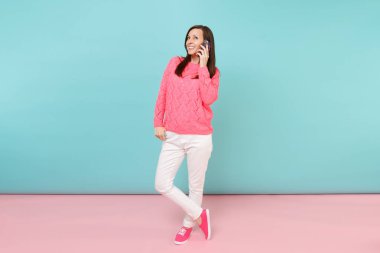 Tam uzunlukta örme gül kazaklı kadın portresi, cep telefonu parlak pembe mavi pastel duvar arka planda Studio izole konuşurken beyaz pantolon. Moda yaşam tarzı kavramı. Kopyalama alanı alay