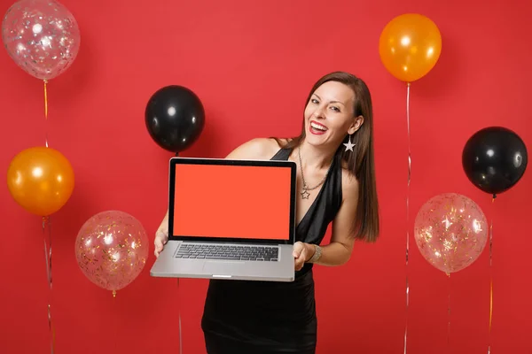 Laughing young woman in black dress celebrating, holding laptop pc computer with blank black empty screen on bright red background air balloons. Happy New Year, birthday mockup holiday party concept