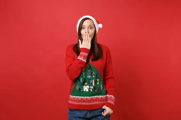 Shocked young Santa girl in knitted sweater, Christmas hat covering mouth with hand isolated on bright red wall background. Happy New Year 2019 celebration holiday party concept. Mock up copy space