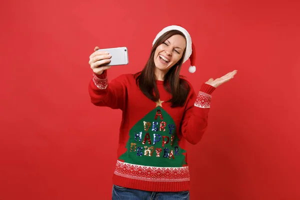 Selfie をやってクリスマス帽子でうれしそうなサンタガールは赤の背景に分離された脇手を指している携帯電話で撮影 幸せな新しい年 2019年お祝いの休日党の概念 コピー スペースをモックアップします — ストック写真