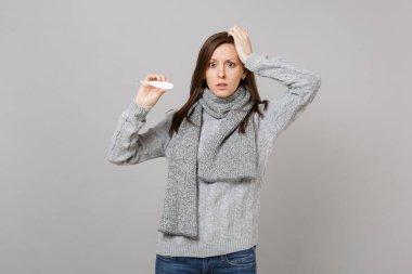 Çapraşık genç kadın eşarp yerine gri süveterli alnına el, gri arka plan üzerinde izole termometre tutun. Sağlıklı yaşam tarzı, hasta hasta hastalık tedavileri, soğuk sezon kavramı. Kopyalama alanı alay