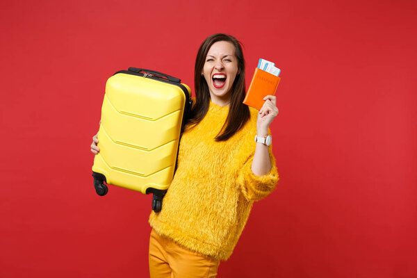 Crazy joyful young woman in yellow fur sweater screaming, holding suitcase, passport boarding pass ticket isolated on red background. People sincere emotions, lifestyle concept. Mock up copy space