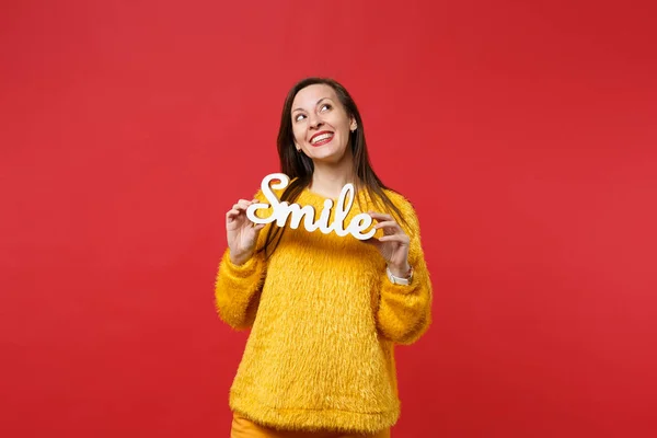 Dreamful young woman in yellow fur sweater looking up, hold wooden word letters smile isolated on bright red wall background in studio. People sincere emotions, lifestyle concept. Mock up copy space