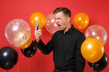 Kızgın genç adam holding, cep telefonuyla kırmızı arka plan hava balonu üzerinde çığlık video görüşmesi siyah klasik gömlek. St. Valentine's gün Happy New Year Doğum günü mockup tatil parti konsept