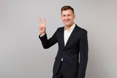 Genç iş adamı zafer işaret Studio gri arka plan üzerinde izole gösteren klasik siyah takım elbise, beyaz gömlek gülümseyen portresi. Başarı kariyer servet iş kavramı. Kopyalama alanı alay