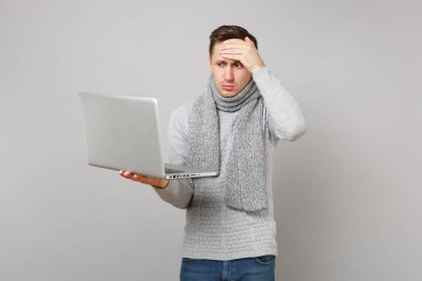 Gri süveterli yorgun genç adam gri arka plan üzerinde izole alnına el koyarak dizüstü pc bilgisayar üzerinde çalışıyor eşarp. Sağlık, online tedavi soğuk sezon kavramı danışmanlık. Kopyalama alanı alay