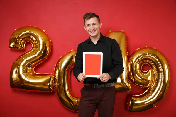 Merry fun young man in black shirt celebrating holiday party hold tablet pc isolated on bright red wall background, golden numbers air balloons studio portrait. Happy New Year 2019 Christmas concept