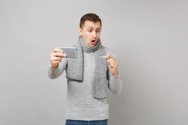 Shocked young man in gray sweater, scarf holding thermometer, doing selfie shot on mobile phone, making video call isolated on grey background. Health ill sick disease treatment, cold season concept