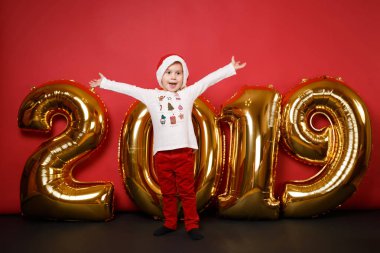 Tatil partisi kutlama Noel şapkalı neşeli Noel Baba Çocuk izole parlak kırmızı duvar arka plan, altın parlak parıltı numaraları hava balonları, tam uzunlukta stüdyo portre. Mutlu yeni yıl 2019 kavramı