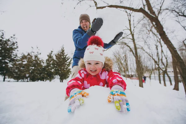 Cheerful family dad in winter warm clothes holding big snowball, little girl playing with snow in park or forest outdoors. Winter fun, leisure on holidays. Love relationship family lifestyle concept