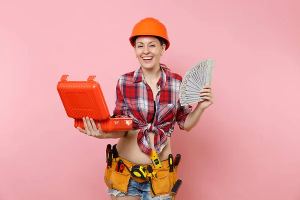 Strong woman in orange helmet, plaid shirt, denim shorts, kit tools belt full of instruments, toolbox, lots cash money isolated on pink background. Female in male work. Renovation occupation concept