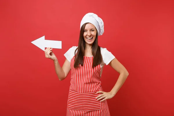 Housewife female chef cook or baker in striped apron, white t-shirt, toque chefs hat isolated on red wall background. Smiling fun housekeeper woman showing side with arrow. Mock up copy space concept