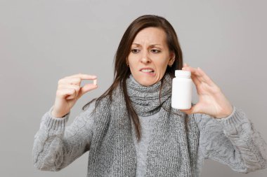 Gri süveterli ilaç tablet aspirin hapı şişe gri arka plan üzerinde izole holding dudak ısırma eşarp şaşkın genç kadın. Sağlıklı yaşam hasta hasta hastalık tedavi soğuk sezon kavramı