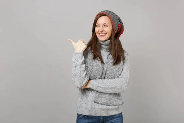 灰色セーター 帽子マフラー見ると 陽気な若い女性は 灰色の背景に分離親指の脇を指しています 健全なライフ スタイル の心から感情 寒い季節の概念 コピー スペースをモックアップします — ストック写真