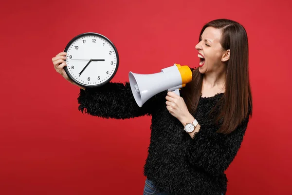 Nervous young woman in black fur sweater screaming on megaphone looking on round clock isolated on bright red wall background in studio. People sincere emotions lifestyle concept. Time is running out