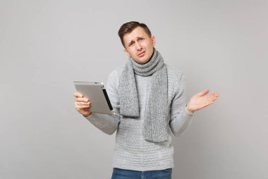 Upset young man in gray sweater, scarf using tablet pc computer, spreading hands isolated on grey background. Healthy lifestyle, online treatment consulting, cold season concept. Mock up copy space