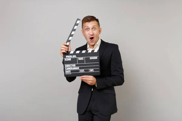 Excited business man keeping mouth wide open, looking surprised hold classic black film making clapperboard isolated on grey background. Achievement career wealth business concept. Mock up copy space