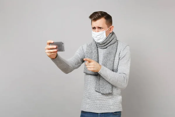 Preoccupied young man in sweater, scarf sterile face mask making video call with mobile phone pointing index finger isolated on grey background. Health ill sick disease treatment, cold season concept