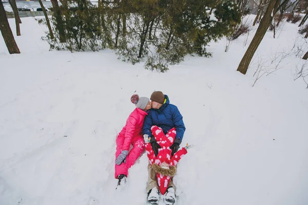 Young family kissing woman and man little girl in warm clothes sitting on snow in park or forest outdoors. Winter fun, leisure on holidays. Love childhood relationship family people lifestyle concept