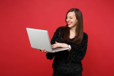 Parlak kırmızı duvar arka planda Studio izole dizüstü pc bilgisayar üzerinde çalışan siyah kürk kazaklı gülen genç kadın portresi. İnsanlar samimi duygular, yaşam kavramı. Kopyalama alanı alay
