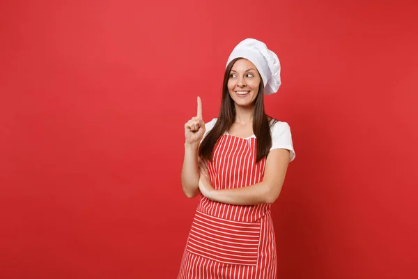 Housewife female chef cook or baker in striped apron, white t-shirt, toque chefs hat isolated on red wall background. Beautiful housekeeper woman pointing index finger up. Mock up copy space concept