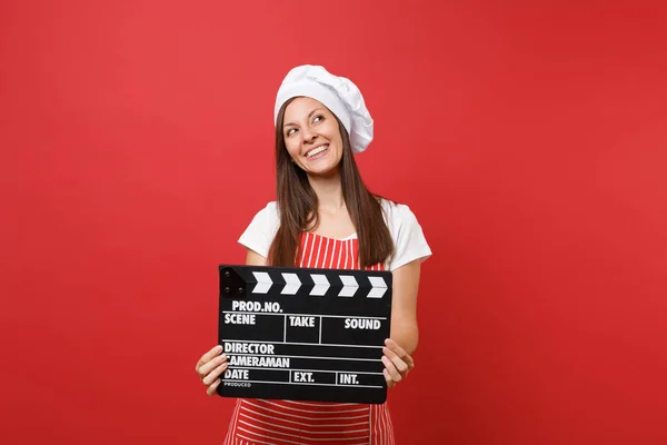 Housewife female chef cook or baker in striped apron, white t-shirt, toque chefs hat isolated on red wall background. Woman holding classic black film making clapperboard. Mock up copy space concept