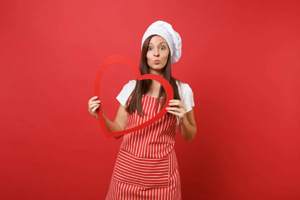 Housewife female chef cook or baker in striped apron, white t-shirt, toque chefs hat isolated on red wall background. Smiling housekeeper woman holding wooden red heart. Mock up copy space concept