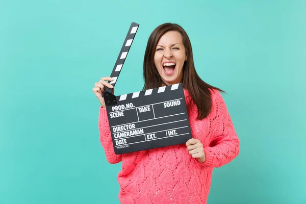 Crazy young woman in knitted pink sweater screaming hold in hand classic black film making clapperboard isolated on blue wall background, studio portrait. People lifestyle concept. Mock up copy space