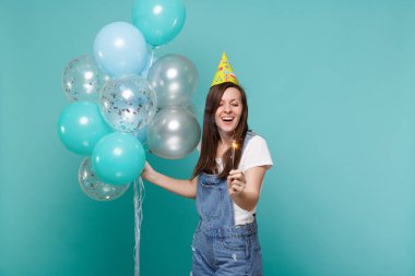 Sevimli gülümseyen genç kadın Doğum günü şapkası holding yanan maytap ve izole mavi turkuaz arka plan üzerinde renkli hava balonları ile kutluyor. Doğum günü tatil partisi, insanların duyguları kavramı