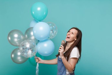 Denim giysi ararken şarkı mikrofon, tutun renkli hava balonları kutluyor mavi turkuaz arka plan üzerinde izole şarkı çekici genç kadın. Doğum günü tatil partisi, insanların duyguları kavramı