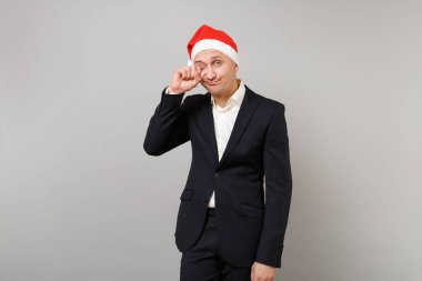 Klasik siyah takım elbise, gri duvar arka plan üzerinde izole gözlerini ovuşturarak Noel şapka içinde uykulu genç iş adamı. Başarı kariyer servet iş. Mutlu yeni yıl 2019 kutlama tatil parti konsept