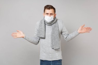 Çapraşık genç adam gri süveterli eşarp steril yüz maskesi gri arka plan üzerinde izole eller yayılıyor. Sağlıklı yaşam tarzı, hasta hasta hastalık tedavileri, soğuk sezon kavramı. Kopyalama alanı alay
