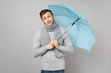 Garip genç adam gri süveterli eşarp gri arka plan üzerinde stüdyo portre izole mavi şemsiye tutan. Sağlıklı moda yaşam tarzı, insanlar samimi duygular, soğuk sezon kavramı. Kopyalama alanı alay