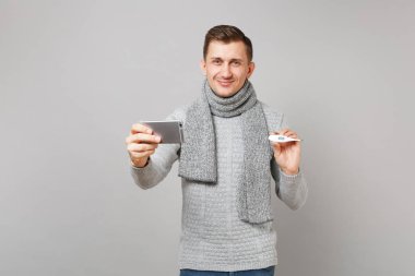 Yakışıklı genç gri süveterli eşarp holding termometre, cep telefonu, ateş selfie gri arka plan üzerinde izole video görüşmesi yapıyor. Sağlık hasta hasta hastalık tedavileri, soğuk sezon kavramı