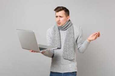 Çapraşık genç adam gri süveterli dizüstü pc bilgisayar üzerinde çalışıyor gri arka plan üzerinde izole termometre tutan eşarp. Sağlık, online tedavi danışmanlık, soğuk sezon kavramı. Kopyalama alanı alay