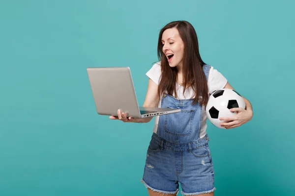 Cheerful young woman football fan holding soccer ball, using laptop pc computer isolated on blue turquoise wall background. People emotions, sport family leisure lifestyle concept. Mock up copy space
