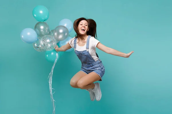 Portrait of pretty funny young woman in denim clothes jumping high celebrating and holding colorful air balloons isolated on blue turquoise background. Birthday holiday party, people emotions concept