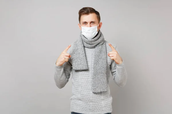 Young man in gray sweater scarf pointing index fingers on sterile face mask isolated on grey background in studio. Healthy lifestyle ill sick disease treatment cold season concept. Mock up copy space