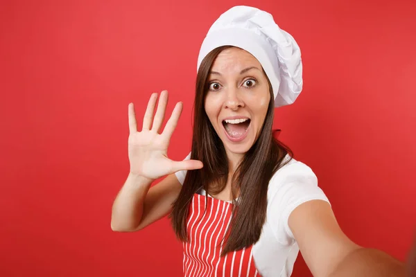 Housewife female chef cook or baker in striped apron, white t-shirt, toque chefs hat isolated on red wall background. Close up housekeeper woman doing taking selfie shot. Mock up copy space concept
