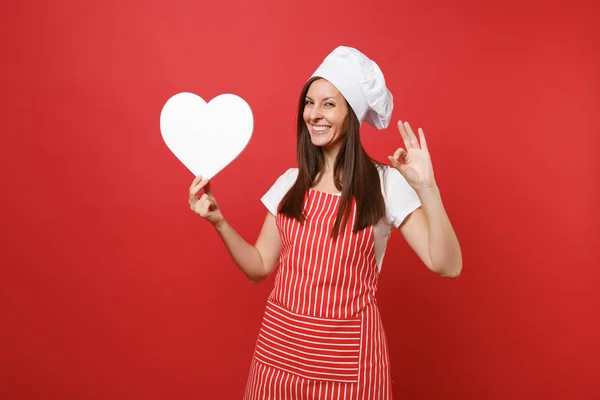 Housewife female chef cook or baker in striped apron, white t-shirt, toque chefs hat isolated on red wall background. Smiling housekeeper woman hold blank heart workspace. Mock up copy space concept