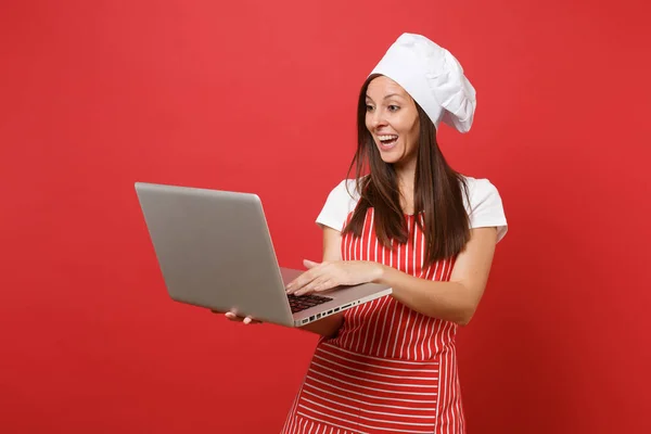 Housewife female chef cook or baker in striped apron, white t-shirt, toque chefs hat isolated on red wall background. Fun housekeeper woman looking for recipe in laptop pc. Mock up copy space concept