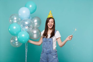 Genç kadın denim giysileri gülüyor, Doğum günü şapka tutun yanan maytap izole mavi turkuaz arka plan üzerinde renkli hava balonları ile kutluyor. Doğum günü tatil partisi, insanların duyguları kavramı