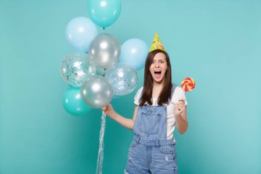 Denim giysiler kızgın çığlık atan kadın, Doğum günü şapka tutun yuvarlak lolipop, izole mavi turkuaz arka plan üzerinde renkli hava balonları ile kutluyor. Doğum günü tatil partisi, insanların duyguları kavramı