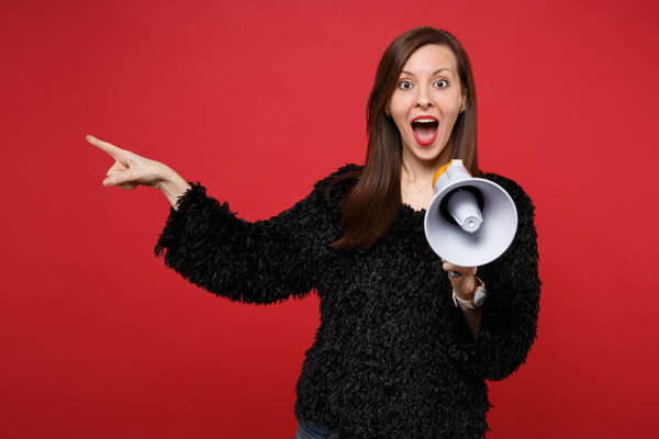 Surprised young woman in black fur sweater with opened mouth pointing index finger aside, holding megaphone isolated on red background. People sincere emotions, lifestyle concept. Mock up copy space