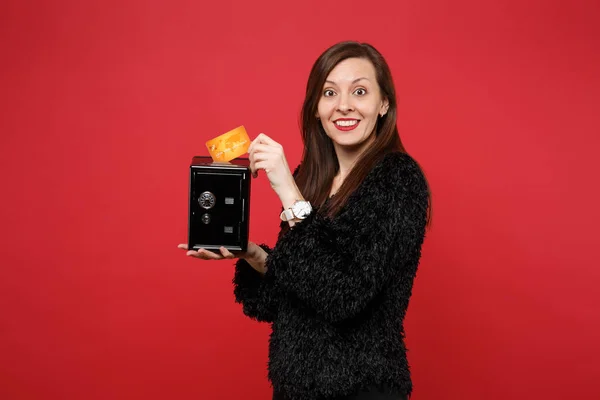 Excited young woman in black fur sweater holding metal bank safe for money accumulation, credt bank card isolated on red background. People sincere emotions, lifestyle concept. Mock up copy space