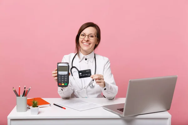 Female doctor sit at desk work on computer with medical document credit card in hospital isolated on pastel pink background. Woman in medical gown glasses stethoscope. Healthcare medicine concept