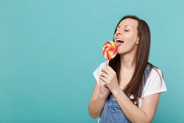 Portrait of cute young woman in denim clothes with closed eyes hold, licking colorful round lollipop isolated on blue turquoise wall background in studio. People lifestyle concept. Mock up copy space
