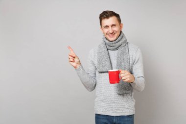 Gri süveterli genç adam gülümsüyor, kırmızı fincan kahve ya da çay, işaret parmağı bir kenara gri arka plan üzerinde izole işaret tutan eşarp. Sağlıklı yaşam tarzı, hasta hasta hastalık tedavileri, soğuk sezon kavramı
